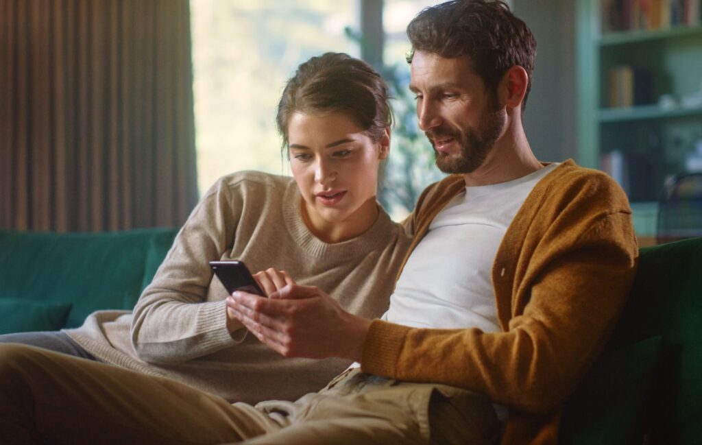 Couple Use Smartphone Device, While Sitting on a Couch in the Cozy Apartment